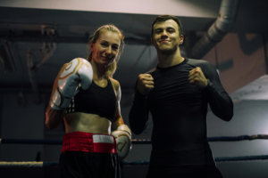 Twee lachende boksers, een vrouw met witte bokshandschoenen en een man in zwarte sportkleding poseren vriendschappelijk samen in de boksring na een training bij Boxing University Breda.